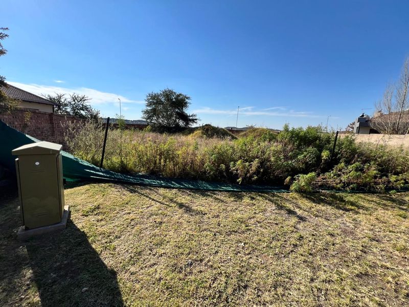 A vacant residential plot featuring overgrown vegetation, a perimeter wall, and an electrical utility box, offering potential for future development.