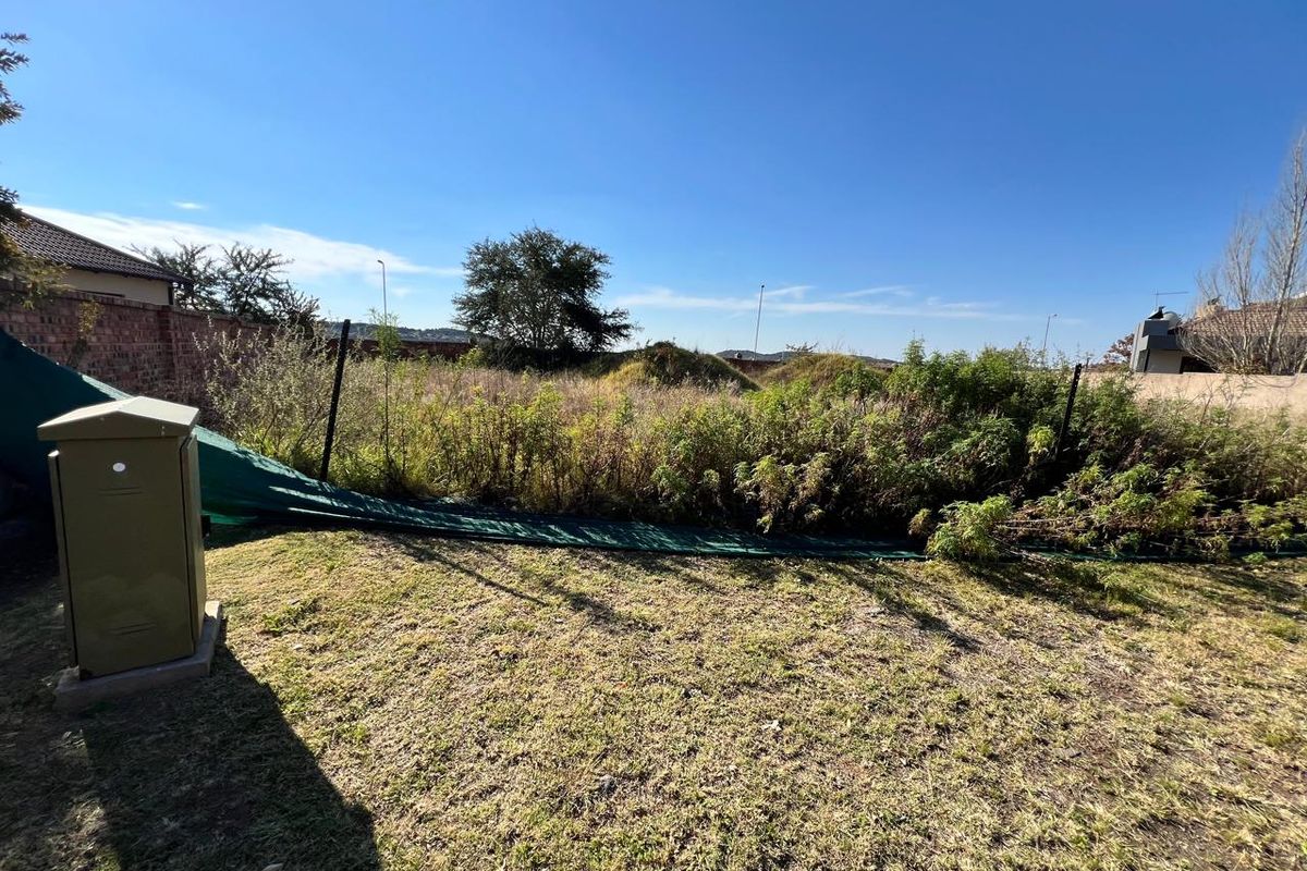 A vacant residential plot featuring overgrown vegetation, a perimeter wall, and an electrical utility box, offering potential for future development.