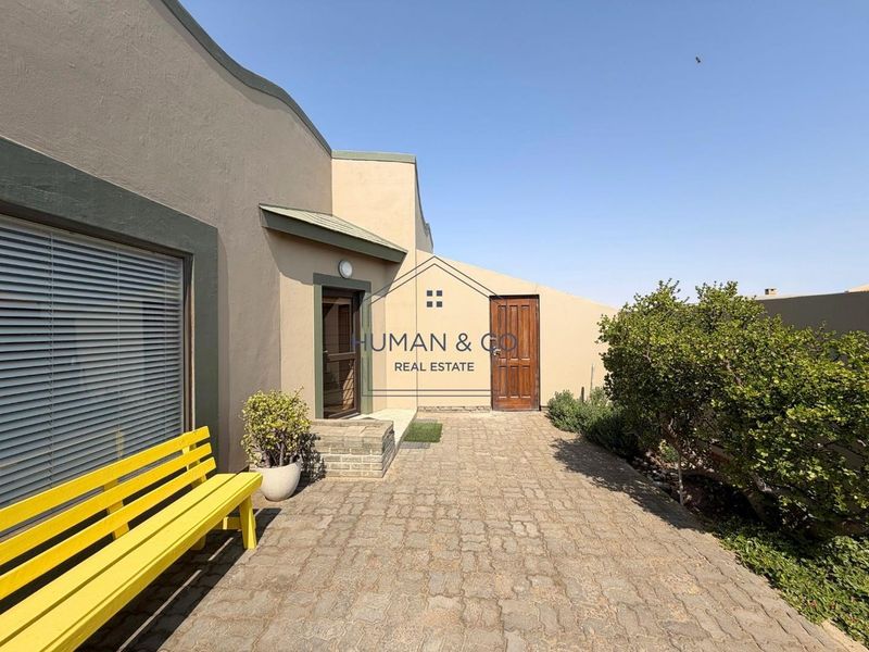 A paved rear patio area featuring a solid timber door, aluminium window frames, and exterior wall lighting, set against a rendered boundary wall.