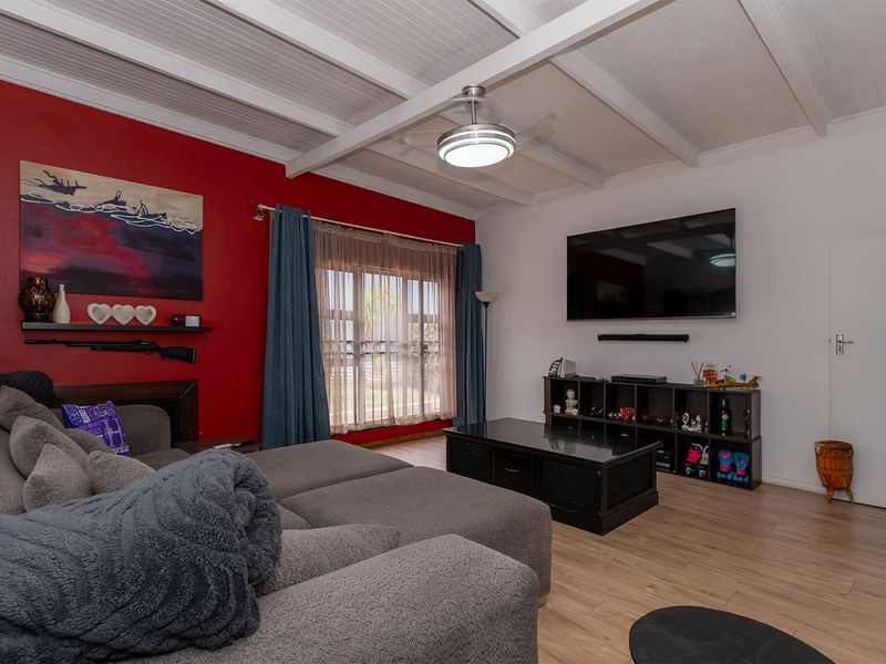 A comfortable living room featuring exposed beam ceilings, a ceiling fan, and laminate flooring, with a bold red accent wall and large window.