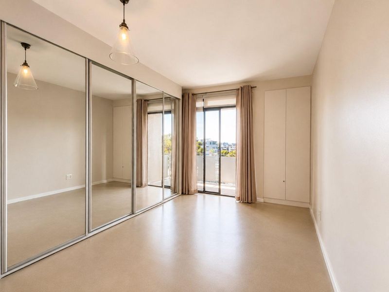A bright, empty bedroom featuring large mirrored sliding wardrobes, built-in storage, and direct access to a private balcony through sliding glass doors.