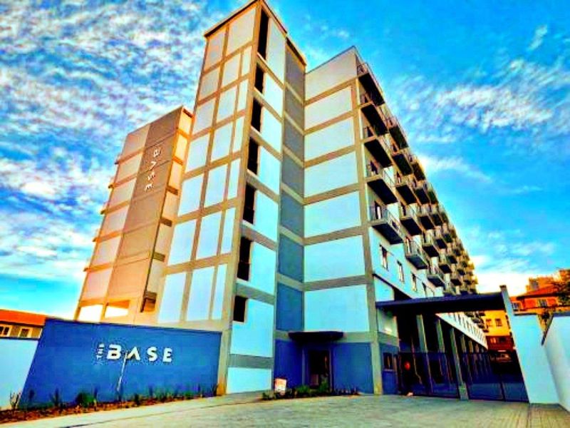 Exterior view of The Base apartment complex featuring a modern multi-story facade with balconies and a paved entrance area.