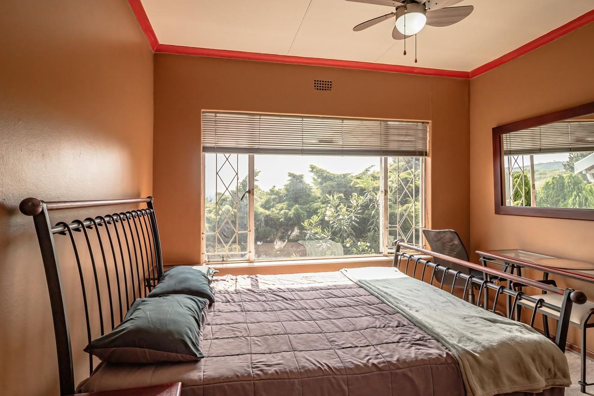 A bedroom featuring a ceiling fan, security bars on the window, and a built-in mirror, offering a view of the surrounding greenery.