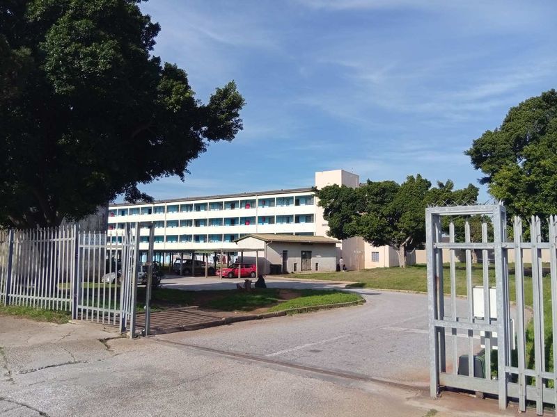 View of the apartment complex entrance featuring a metal security gate, paved driveway, and multi-story residential building.