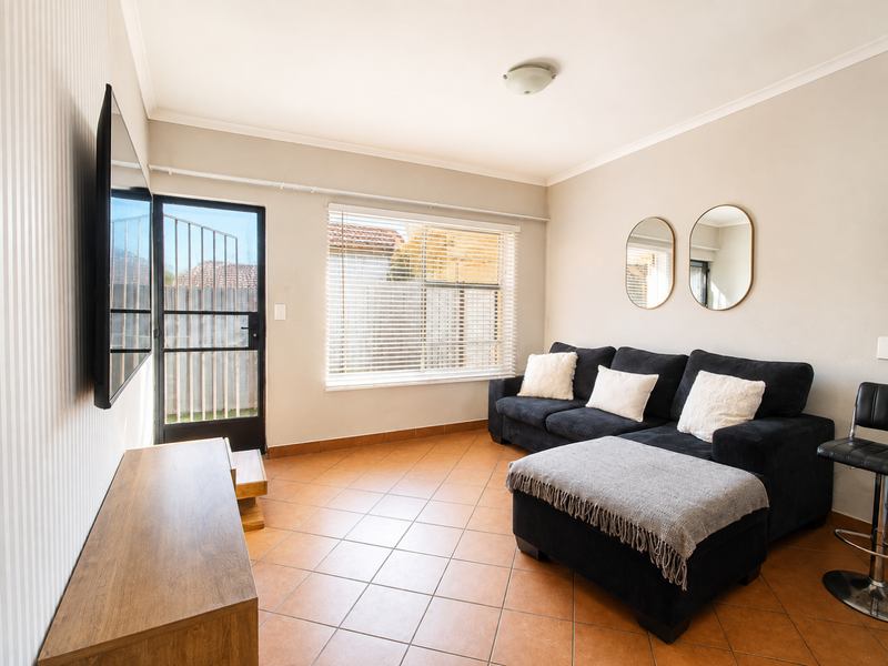 A living room featuring tiled flooring, a security gate leading outdoors, and venetian blinds, providing a functional and well-lit space.