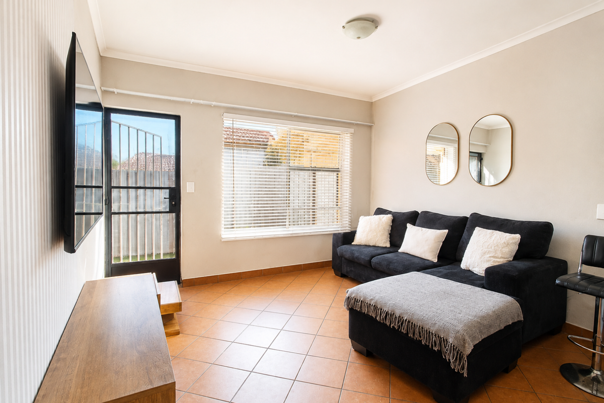 A living room featuring tiled flooring, a security gate leading outdoors, and venetian blinds, providing a functional and well-lit space.