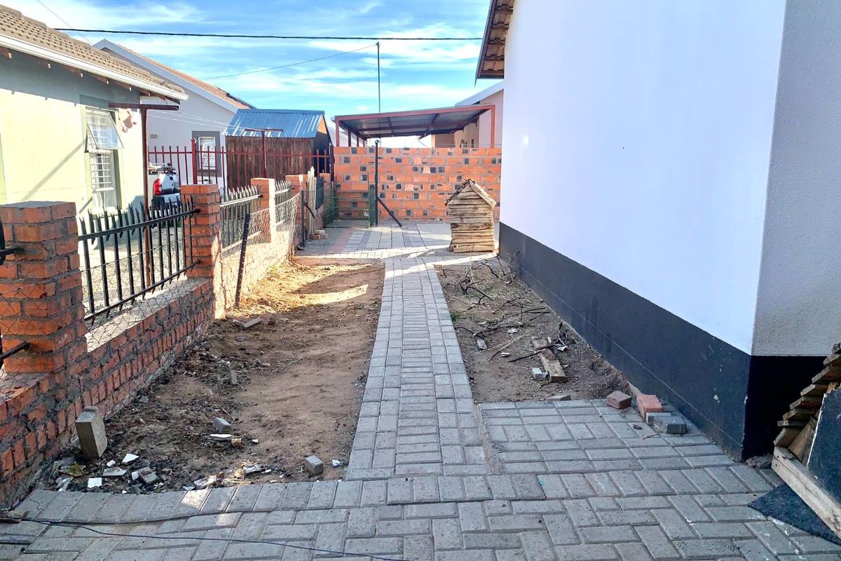 A side exterior pathway featuring paved walkways, brick boundary walls, and unfinished garden beds requiring landscaping.
