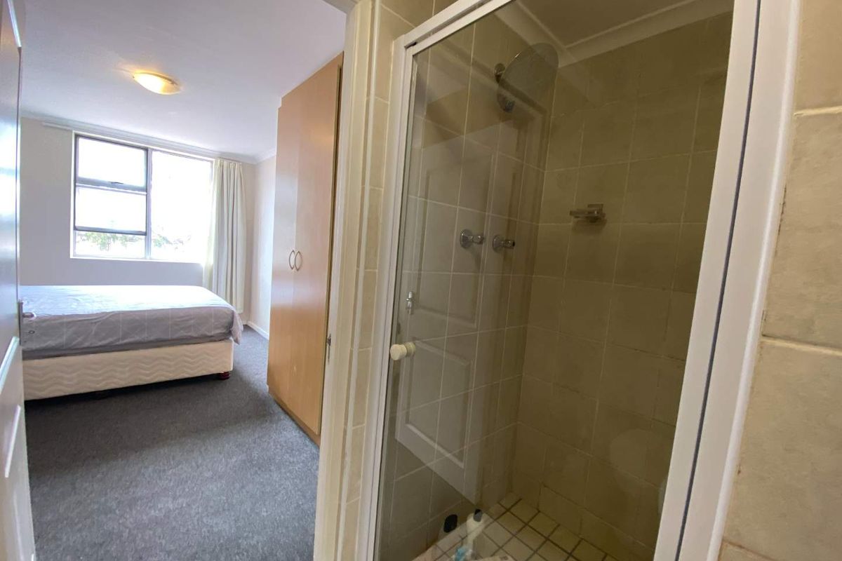 A view from a bathroom into an adjacent bedroom, featuring a glass-enclosed shower with tiled walls and carpeted flooring.