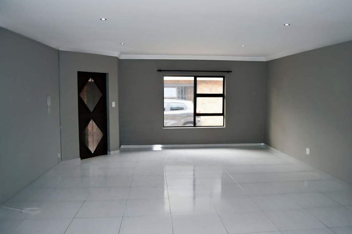 A spacious, empty living area featuring neutral grey walls, polished white floor tiles, recessed ceiling lighting, and a dark-framed window and door.
