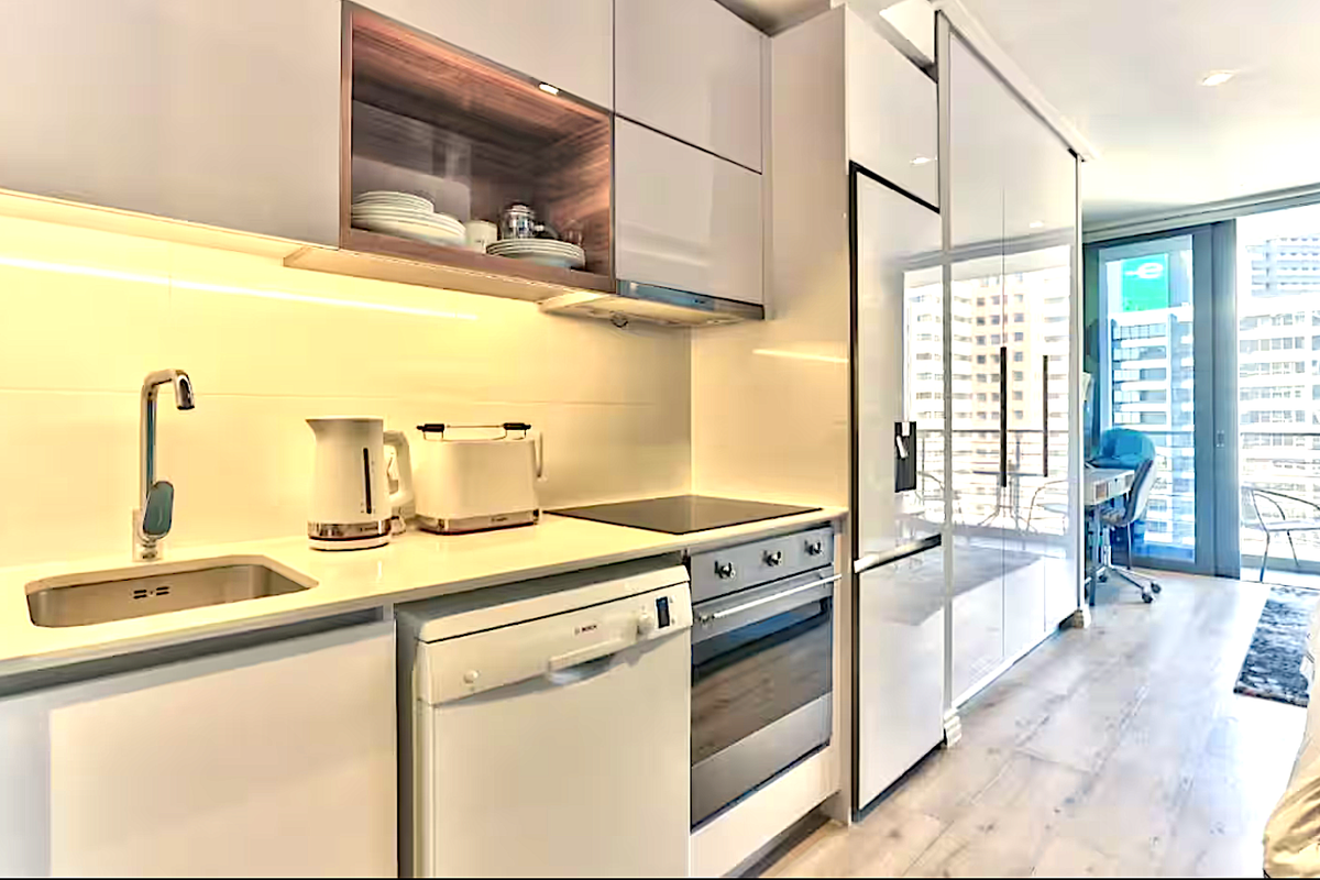 Modern compact kitchen featuring high-gloss cabinetry, integrated appliances, sleek stone countertops, and under-cabinet lighting, seamlessly flowing into the adjacent living area.
