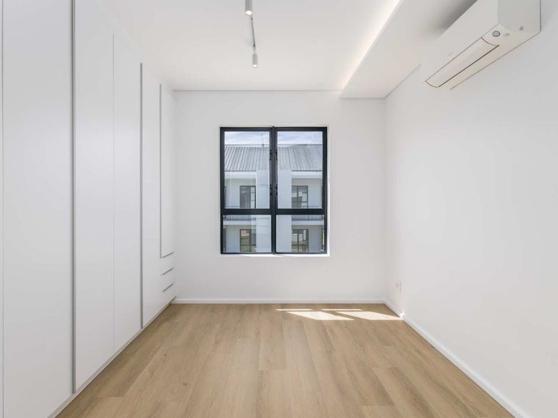 A bright, modern, and empty bedroom featuring light laminate flooring, extensive built-in cupboards, and a wall-mounted air conditioning unit.