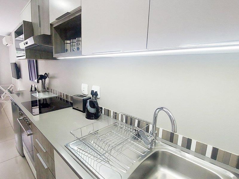 Modern galley-style kitchen featuring sleek cabinetry, integrated appliances, a stainless steel sink, and contemporary tiled backsplash with under-cabinet lighting.