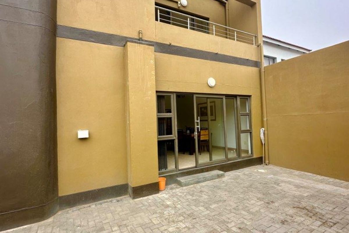 Rear exterior view of a multi-story house featuring a paved courtyard, sliding glass doors, and an upper-level balcony with metal railings.