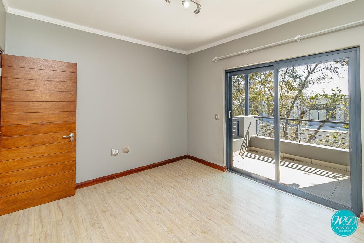 A bright, empty bedroom featuring light laminate flooring, a solid wooden door, and a sliding glass door providing access to a private balcony.