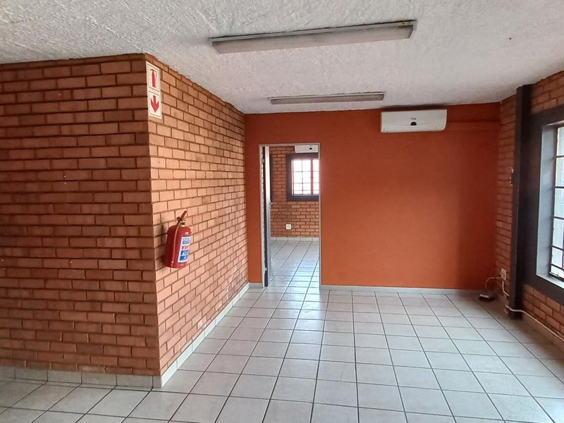 An open-plan industrial office space featuring exposed brick walls, tiled flooring, fluorescent lighting, and a wall-mounted air conditioning unit.