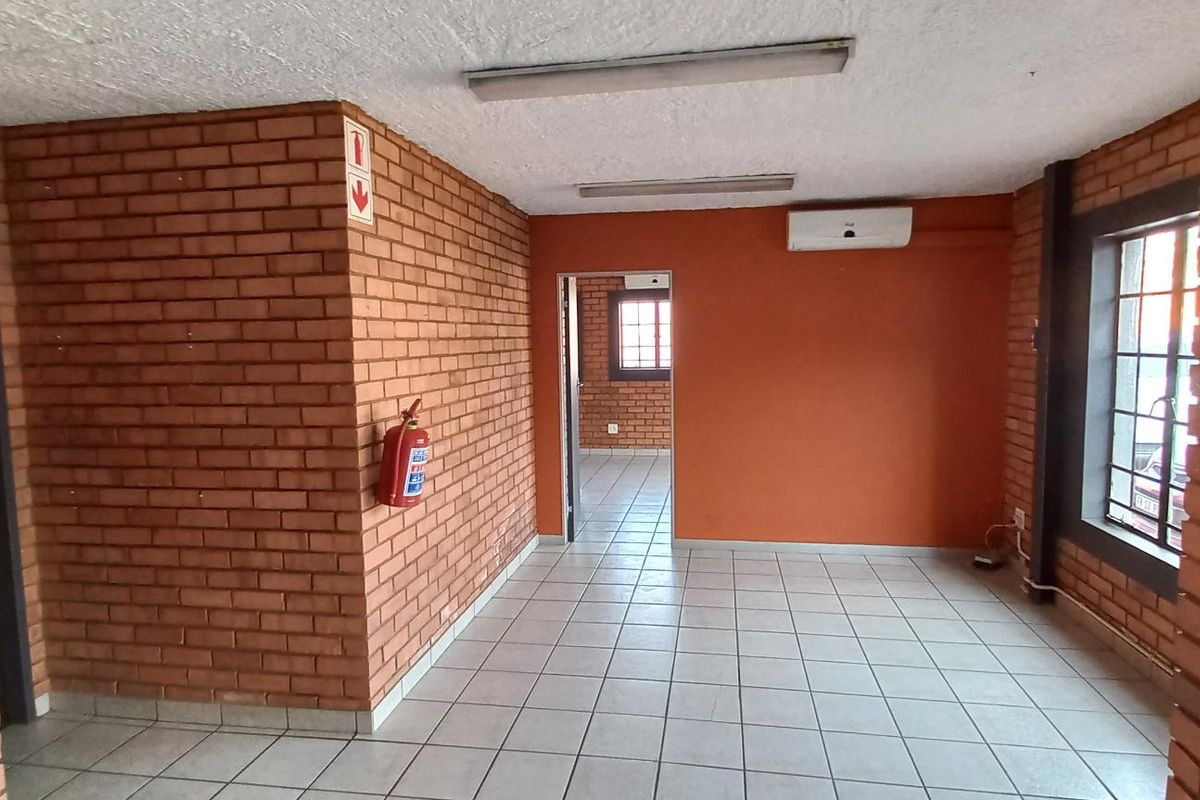 An open-plan industrial office space featuring exposed brick walls, tiled flooring, fluorescent lighting, and a wall-mounted air conditioning unit.