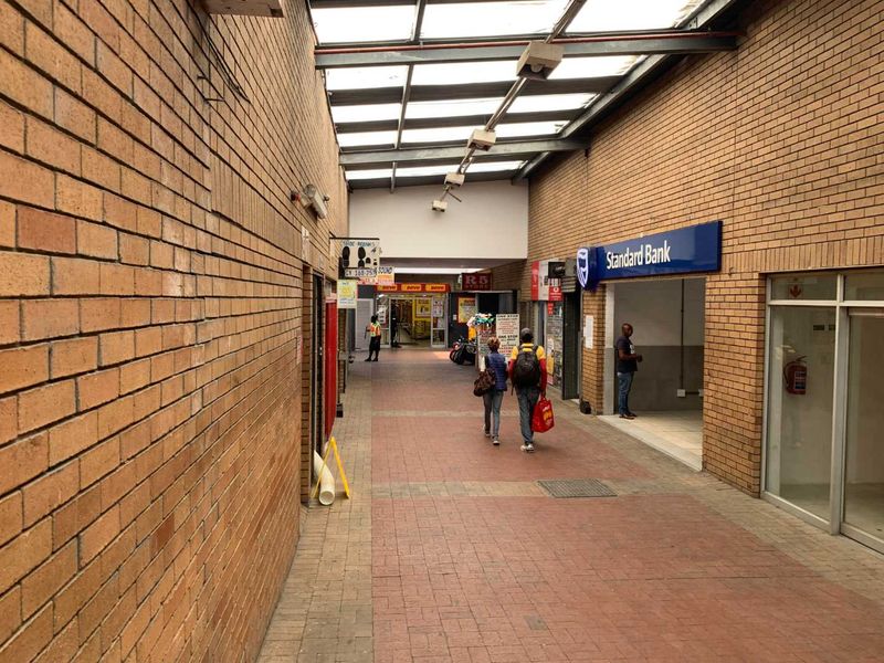 A covered retail walkway featuring exposed brick walls, paved flooring, and a translucent roof structure providing natural light to the commercial corridor.