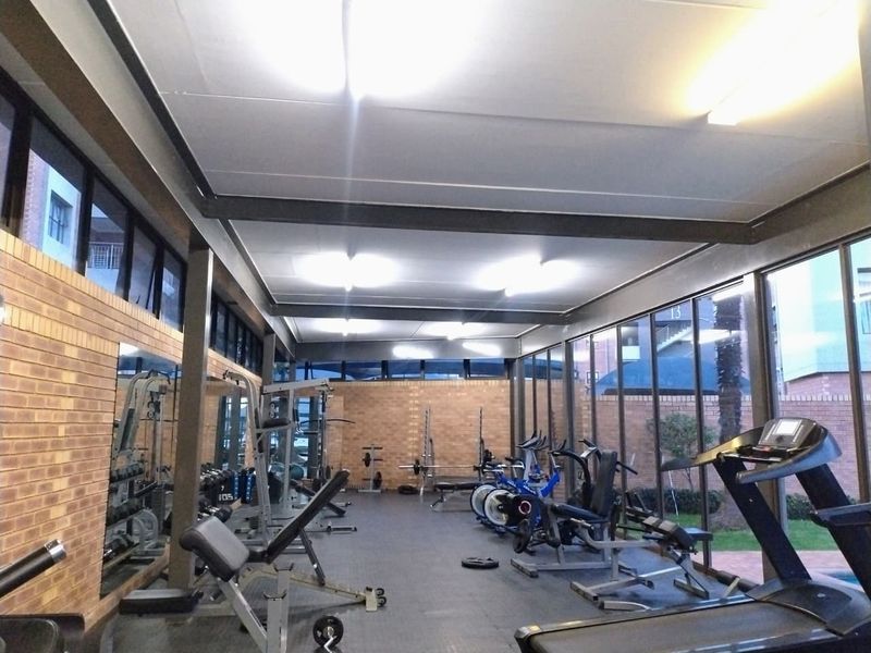 A well-lit communal gym facility featuring exposed steel structural beams, face brick walls, and extensive floor-to-ceiling windows overlooking the exterior grounds.