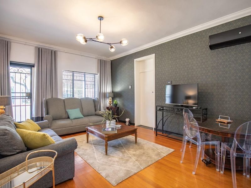 A well-maintained living room featuring hardwood flooring, crown molding, a modern chandelier, and a wall-mounted air conditioning unit.