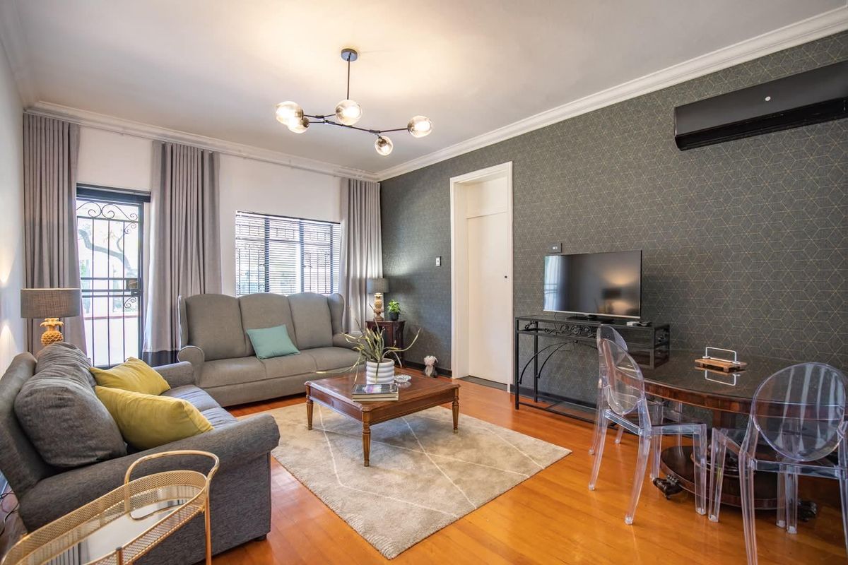 A well-maintained living room featuring hardwood flooring, crown molding, a modern chandelier, and a wall-mounted air conditioning unit.