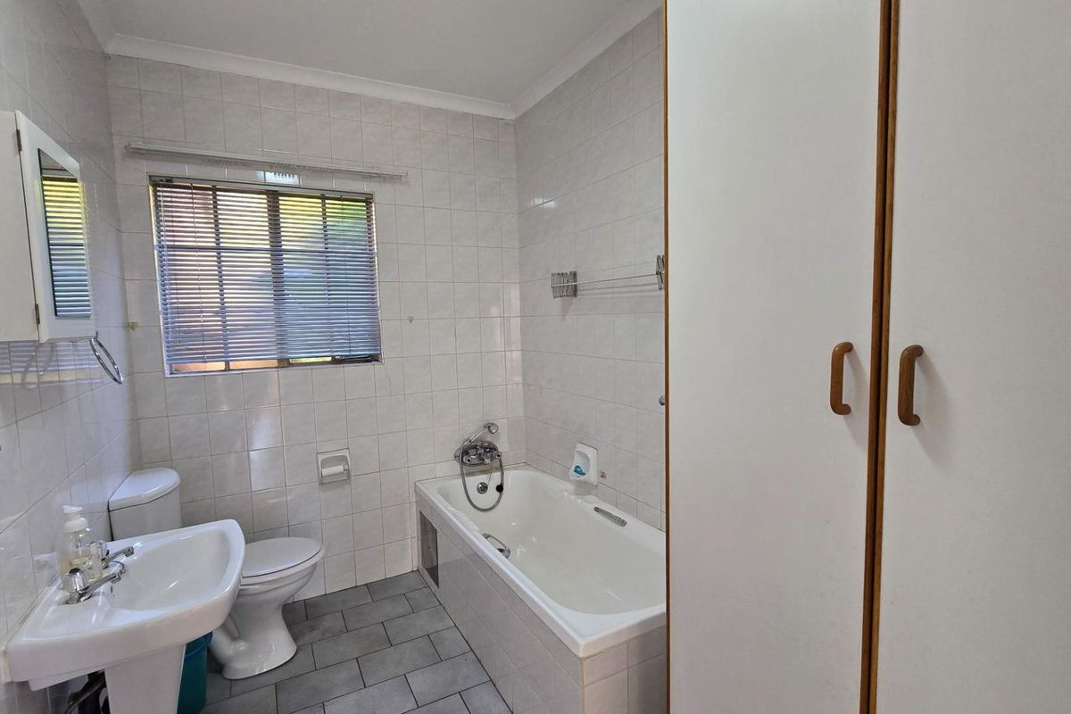 A functional bathroom featuring a built-in bathtub, pedestal basin, toilet, and integrated storage cupboard with neutral wall and floor tiling.