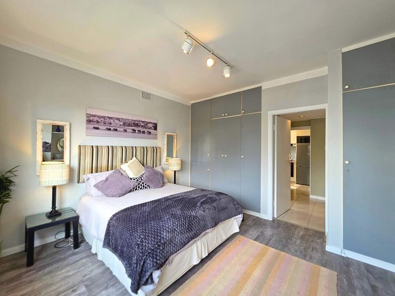 A well-maintained bedroom featuring built-in cupboards, modern track lighting, and durable laminate flooring, with an adjoining view into the hallway.