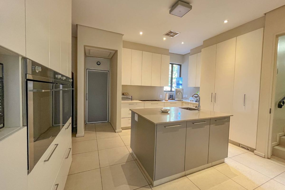 Modern kitchen featuring a central island with a sink, built-in double ovens, ample cabinetry, and neutral tiled flooring.
