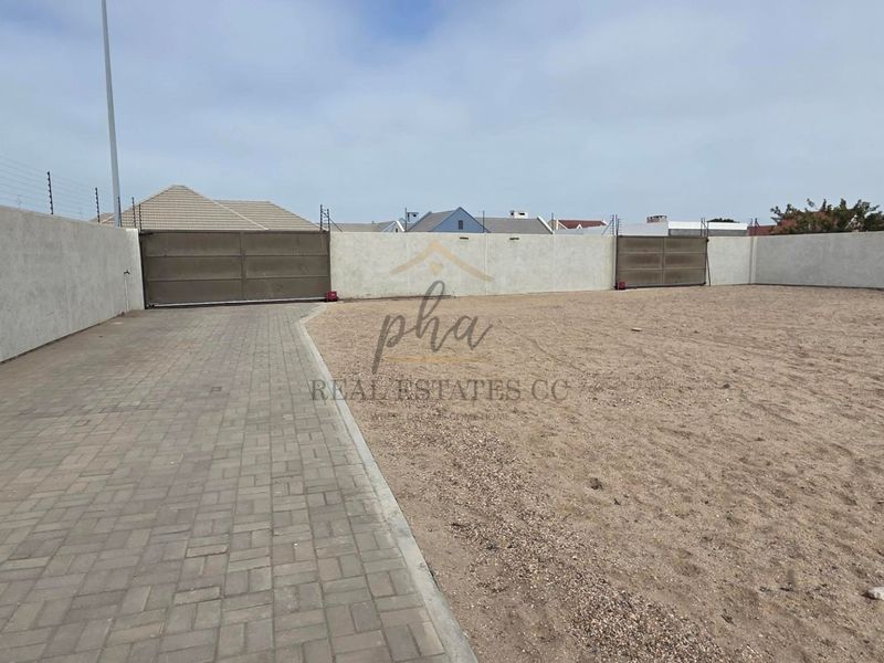 A spacious, secure property entrance featuring a paved driveway, perimeter walling with electric fencing, and large sliding gates.