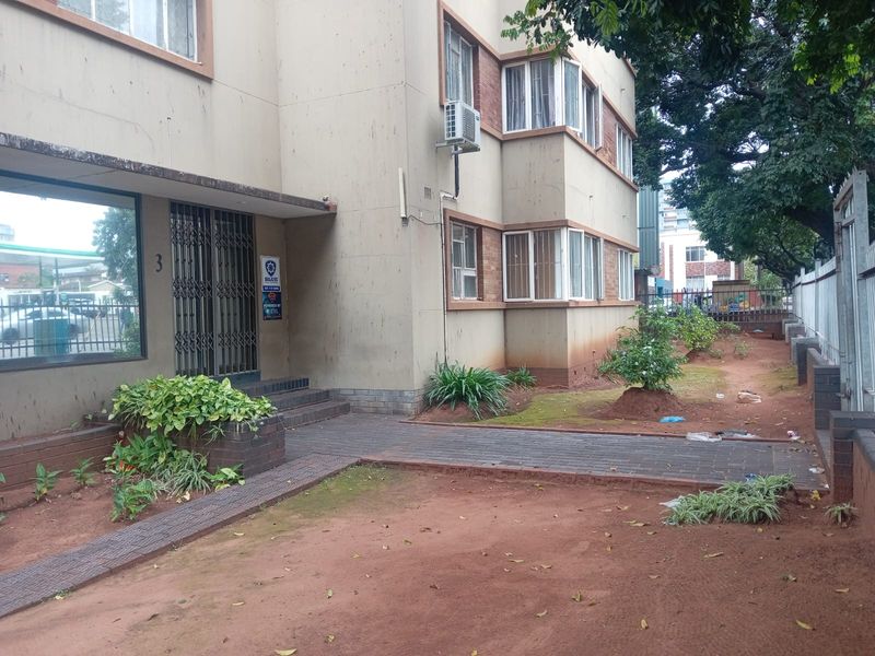 Front exterior view of an apartment building featuring brick paving, security gate entrance, and external air conditioning units with surrounding garden area.