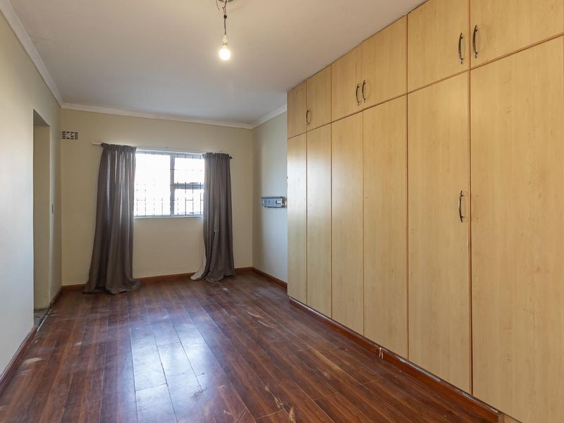 A spacious bedroom featuring extensive built-in wooden cupboards, laminate flooring, and security bars on the window.