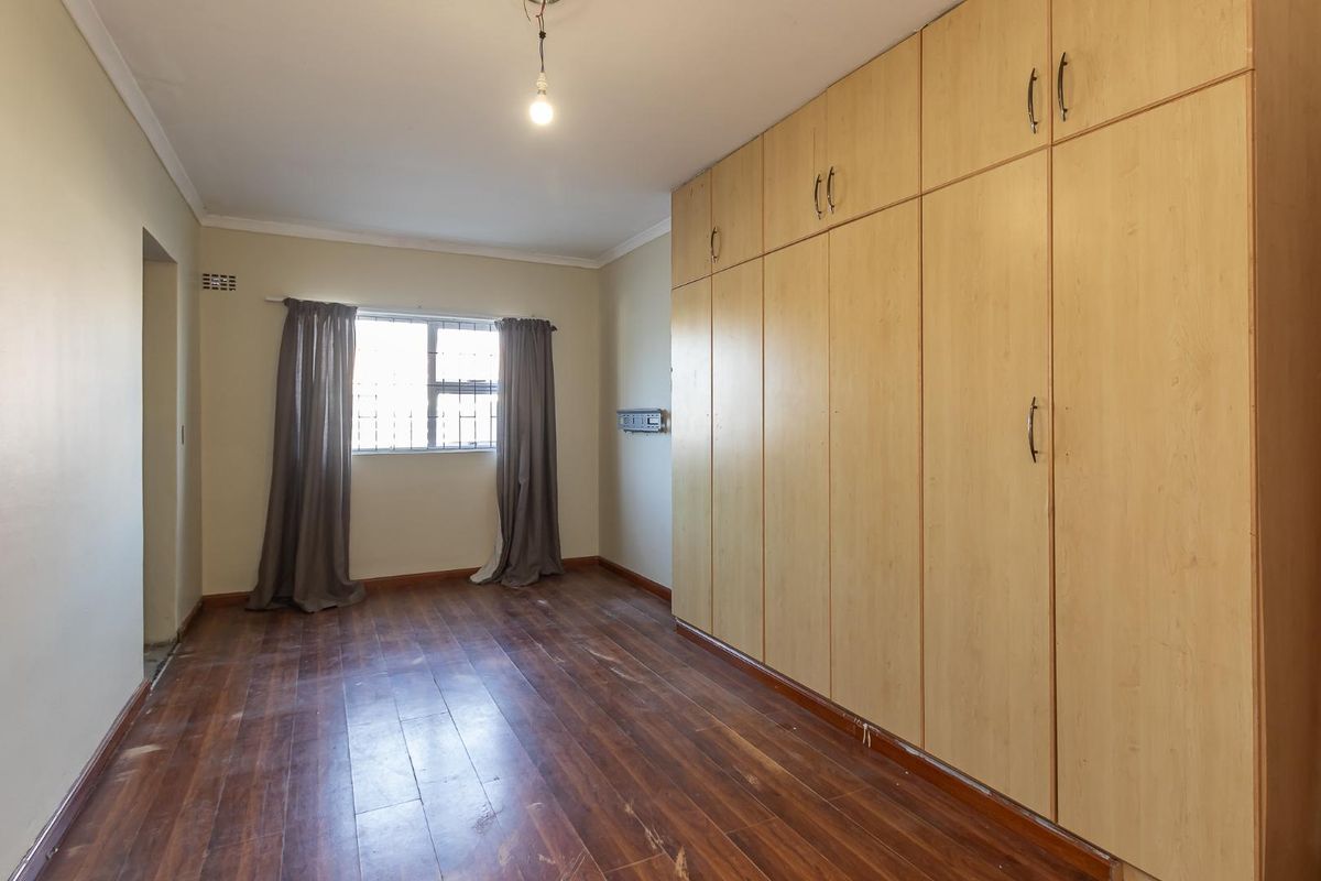 A spacious bedroom featuring extensive built-in wooden cupboards, laminate flooring, and security bars on the window.