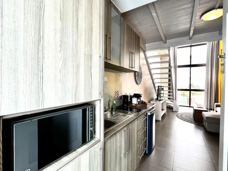 Open-plan apartment living area featuring a modern kitchenette with built-in cabinetry, electric hob, and microwave, leading to a bright space with staircase access.