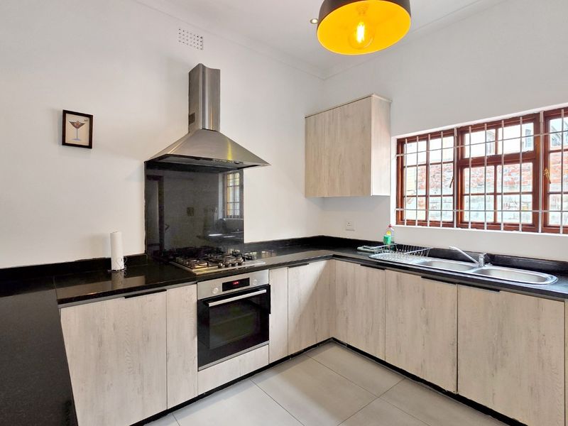 Modern kitchen featuring light wood-grain cabinetry, black granite countertops, stainless steel built-in oven, gas hob, extractor fan, and a double sink with window security bars.