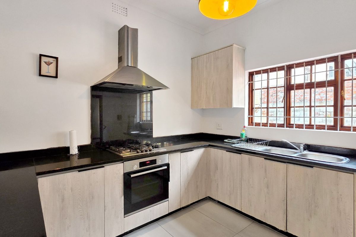 Modern kitchen featuring light wood-grain cabinetry, black granite countertops, stainless steel built-in oven, gas hob, extractor fan, and a double sink with window security bars.