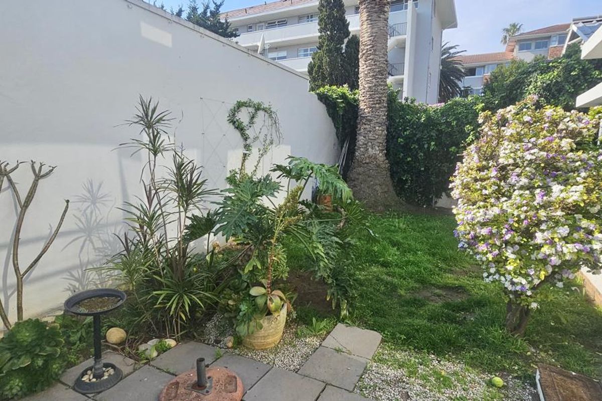 A private rear garden featuring a paved patio area, established greenery, and direct access to the apartment via sliding glass doors.