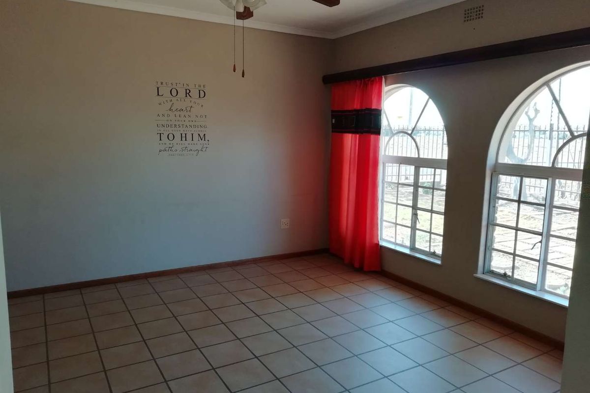 A spacious living room featuring ceramic floor tiles, arched windows with security bars, and a ceiling fan.