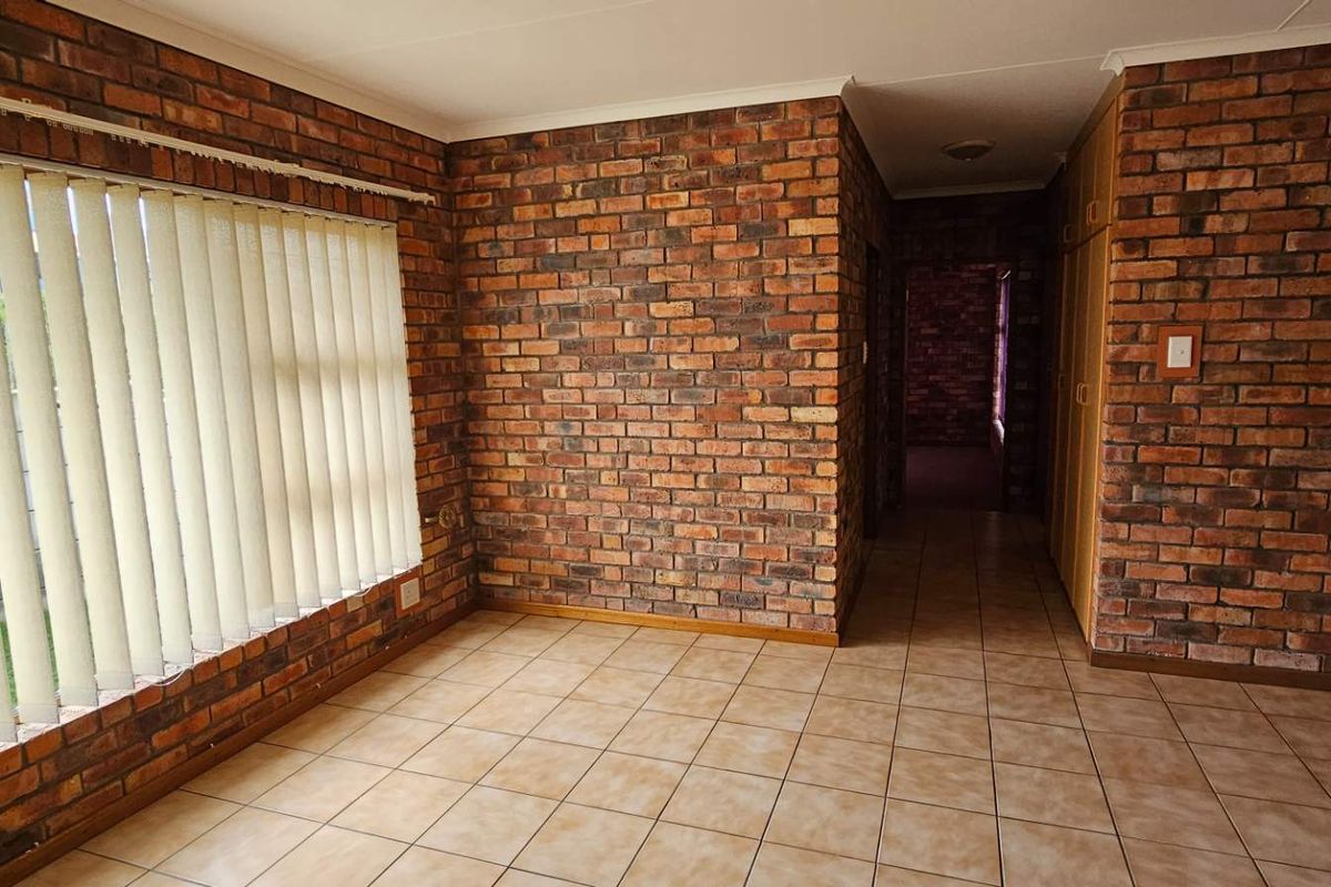 Spacious living area featuring rustic exposed brick walls, neutral ceramic floor tiles, and large windows with vertical blinds, offering a blank canvas for interior design.