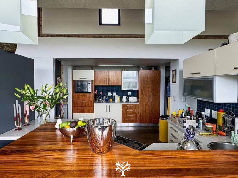 A functional kitchen featuring timber cabinetry, a large wooden breakfast bar, tiled backsplash, and integrated appliances under a high ceiling with a skylight.