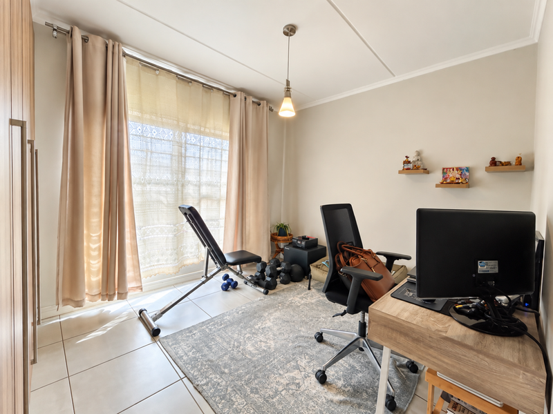 A functional bedroom space featuring tiled flooring, a built-in wardrobe, and natural light through large windows, currently utilized as a home office and gym.