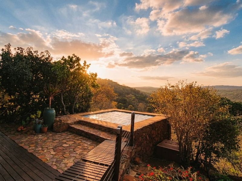 A scenic outdoor plunge pool area featuring stone cladding, timber decking, and expansive elevated views of the surrounding landscape at sunset.