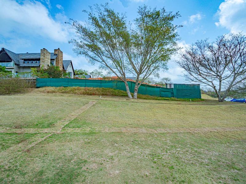 A view of a sloped residential plot featuring mature trees and green privacy screening, with neighboring property visible in the background.