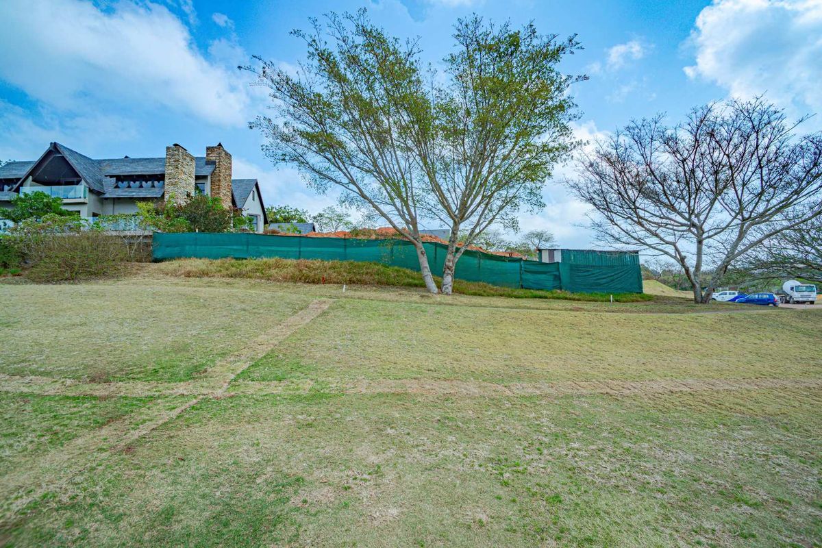 A view of a sloped residential plot featuring mature trees and green privacy screening, with neighboring property visible in the background.