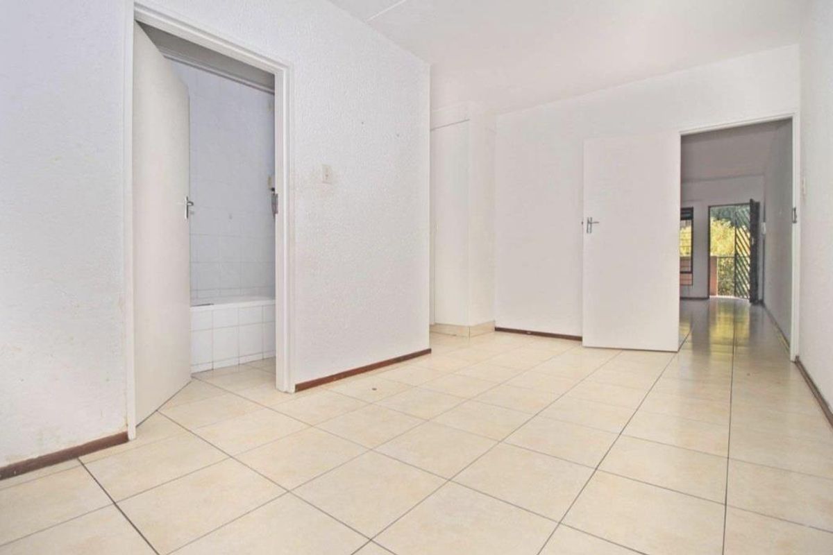 An empty living area featuring tiled flooring and access to an adjoining bathroom and hallway, presenting a blank canvas for renovation.