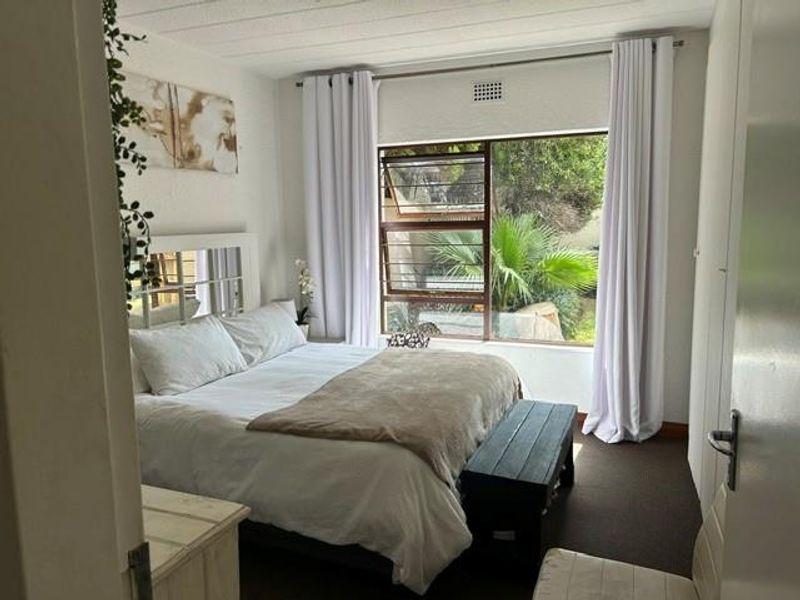 A well-lit bedroom featuring built-in cupboards, carpeted flooring, and a large window with security bars overlooking the garden.