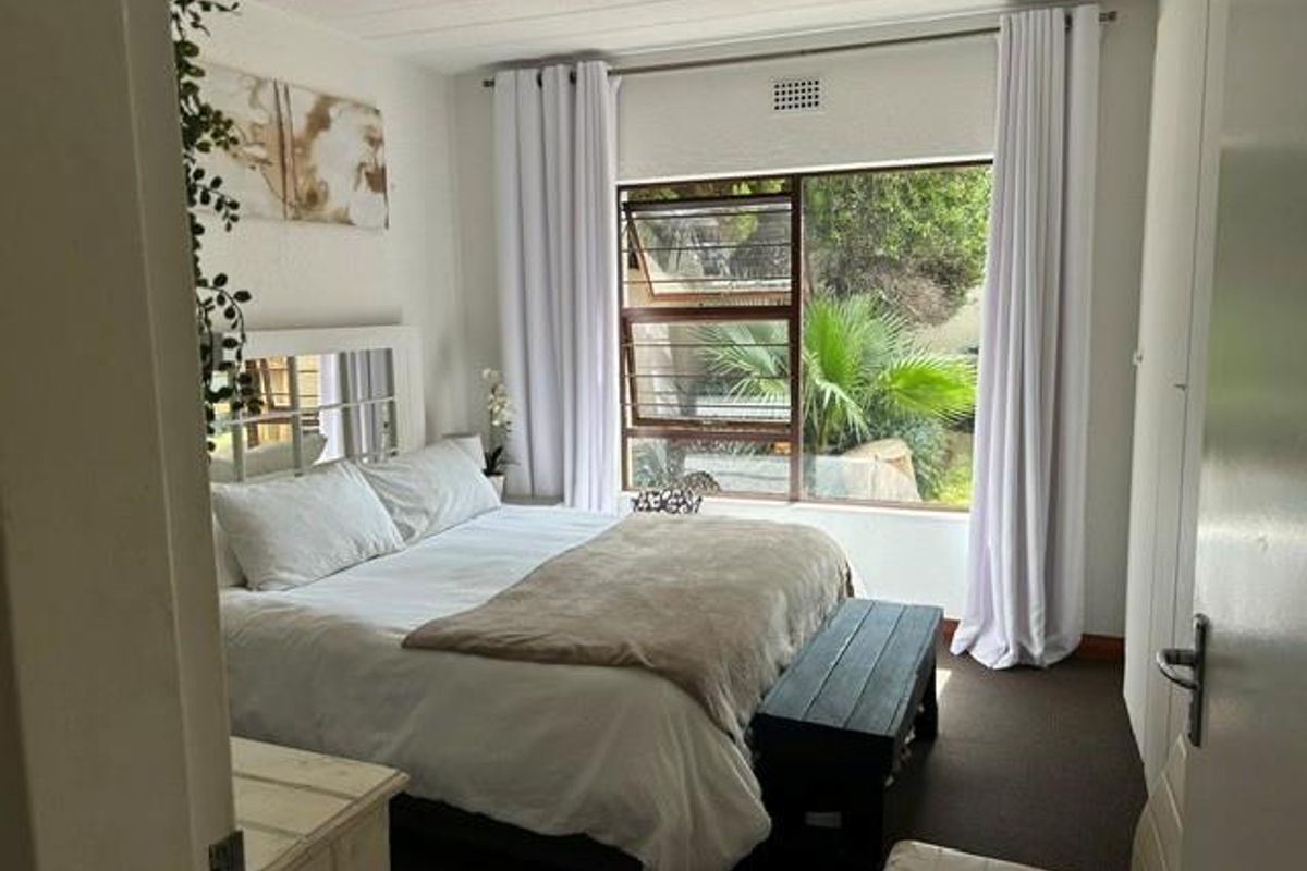 A well-lit bedroom featuring built-in cupboards, carpeted flooring, and a large window with security bars overlooking the garden.