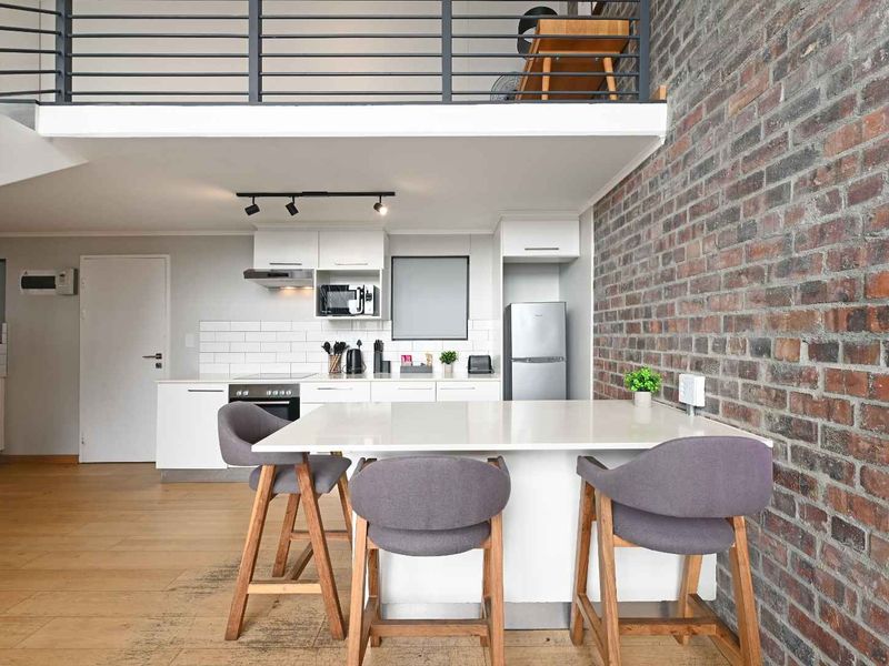 Modern open-plan kitchen featuring a central island, sleek cabinetry, and an exposed brick wall, with a mezzanine level overlooking the living space.