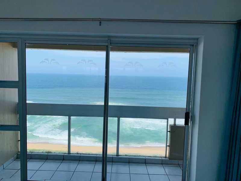 A clear view of the ocean and beach from an apartment balcony, featuring a sliding glass door and tiled outdoor flooring.