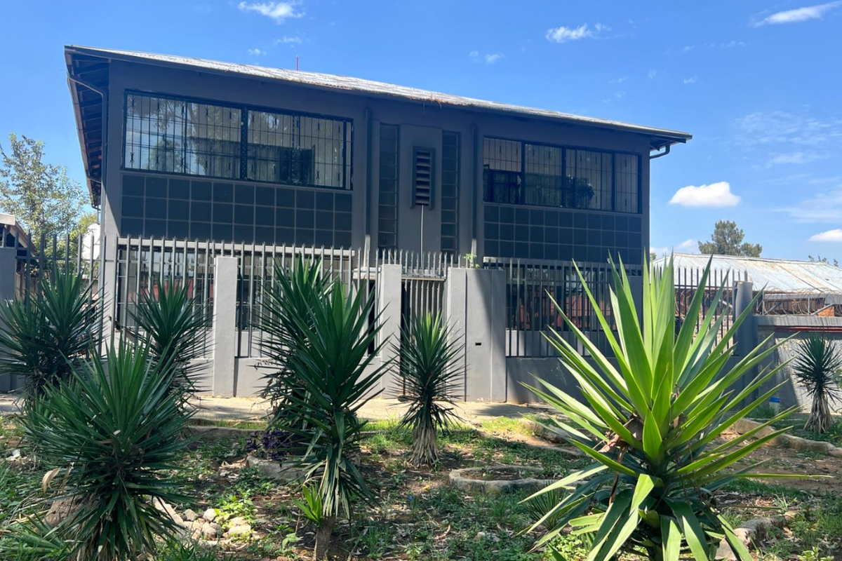 Rear view of a two-story commercial building featuring a dark grey facade, security bars on windows, and a metal roof, set within a landscaped garden.
