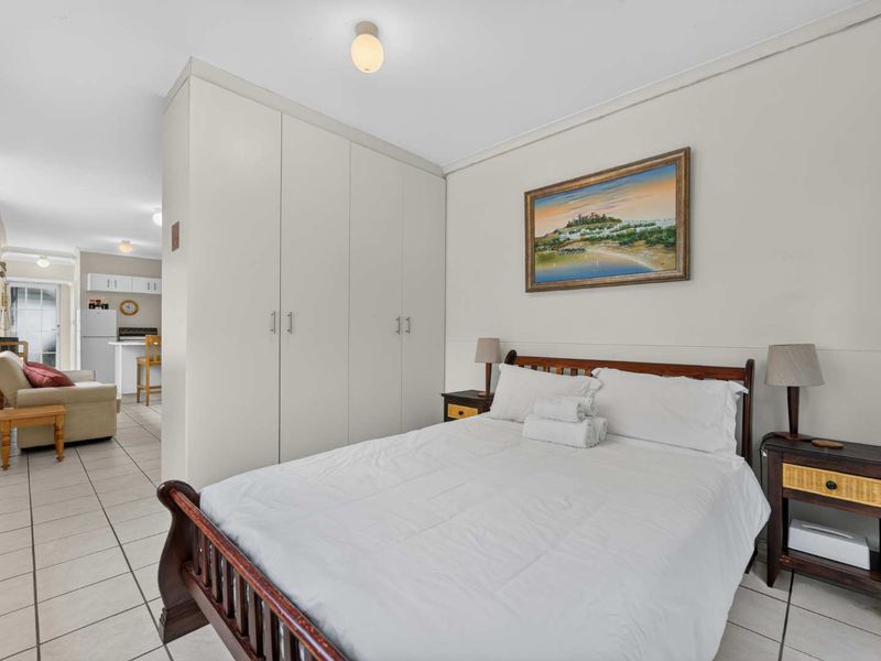 A bedroom featuring built-in wardrobes and tiled flooring, with an open-plan layout leading into the adjacent living and kitchen area.
