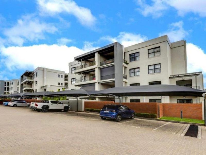 Exterior view of a modern multi-story apartment complex featuring paved parking areas with shade cloth carports and brick boundary walls.